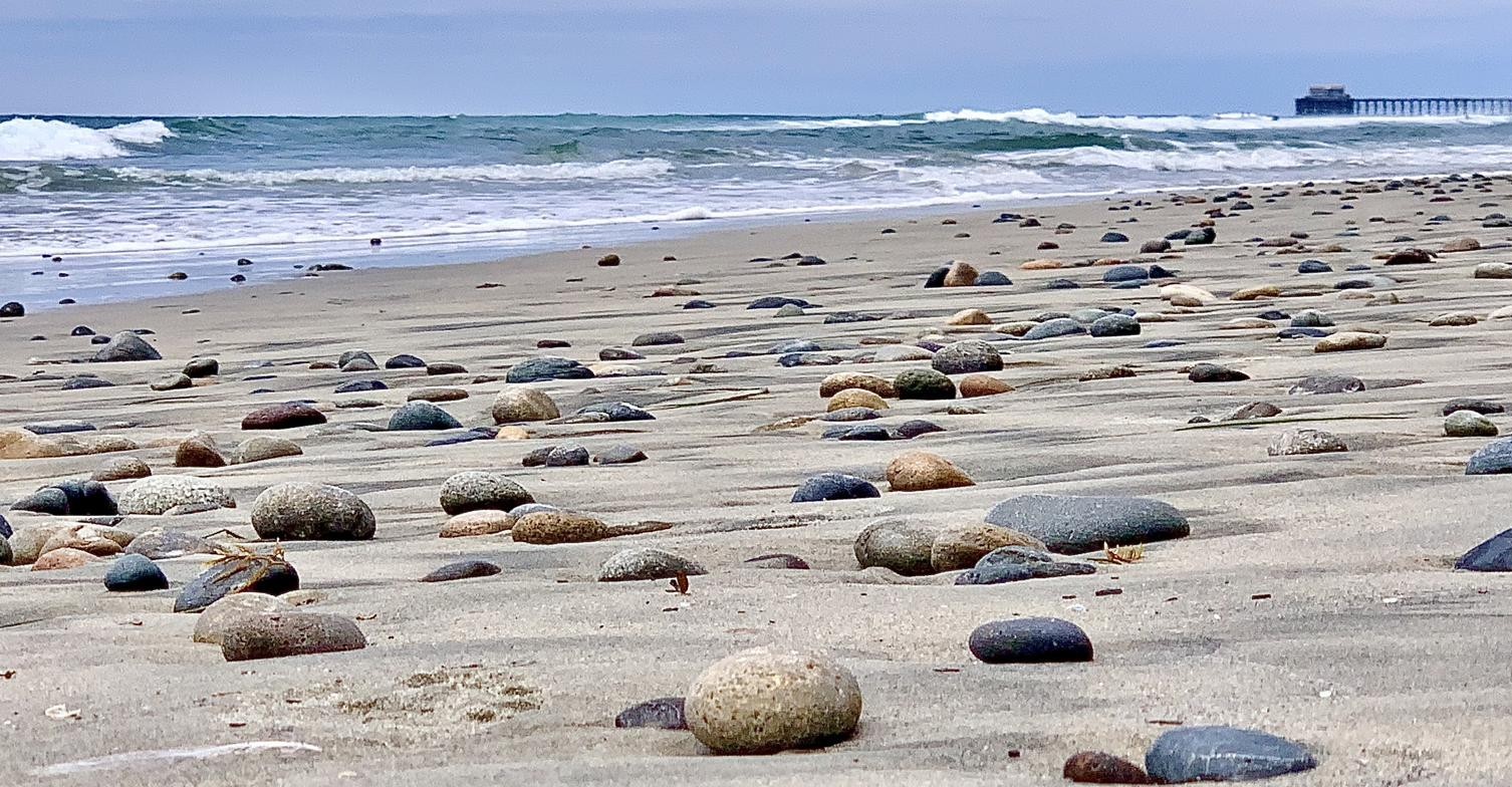 The Origin — beach scattered with stones, waves and pier in the distance